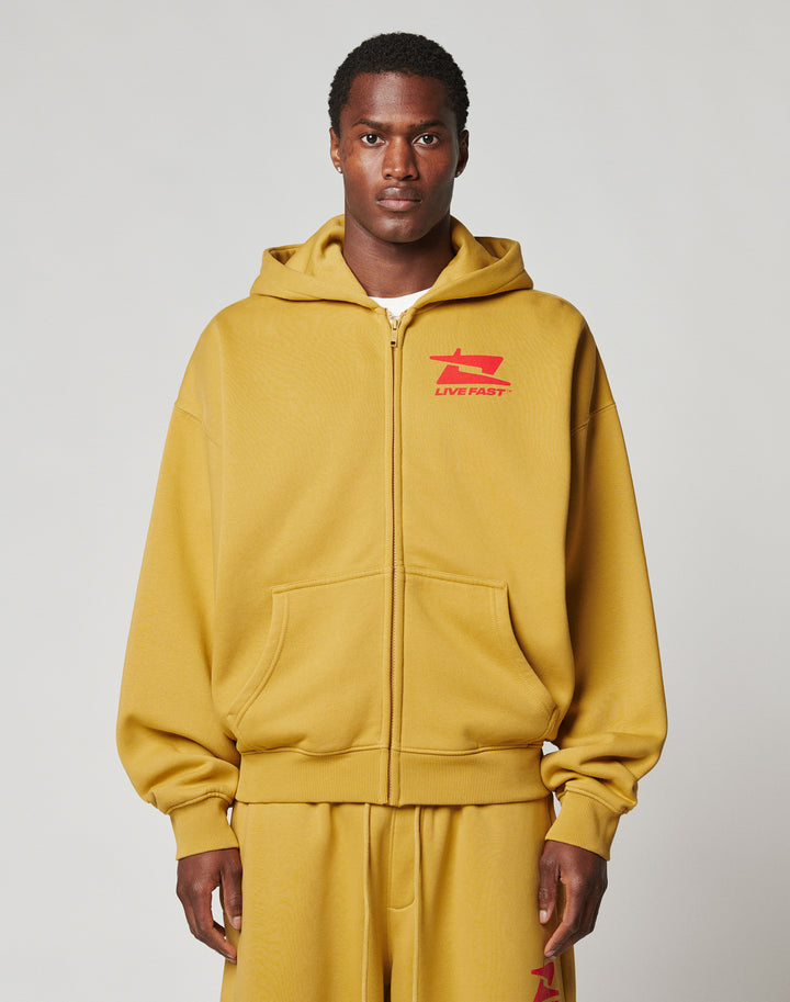 A man faces forward against a light gray background, wearing the LIVE FAST DIE YOUNG Live Fast Sweatsuit Jacket in mustard yellow with a bold LIVE FAST front print and red chest graphic, paired with matching oversized sweatpants. | model