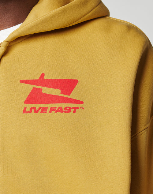 Close-up of the Live Fast Sweatsuit Jacket by LIVE FAST DIE YOUNG in mustard yellow, oversize fit, with a red front graphic and LIVE FAST text on the chest. Visible hood and part of a white shirt underneath.