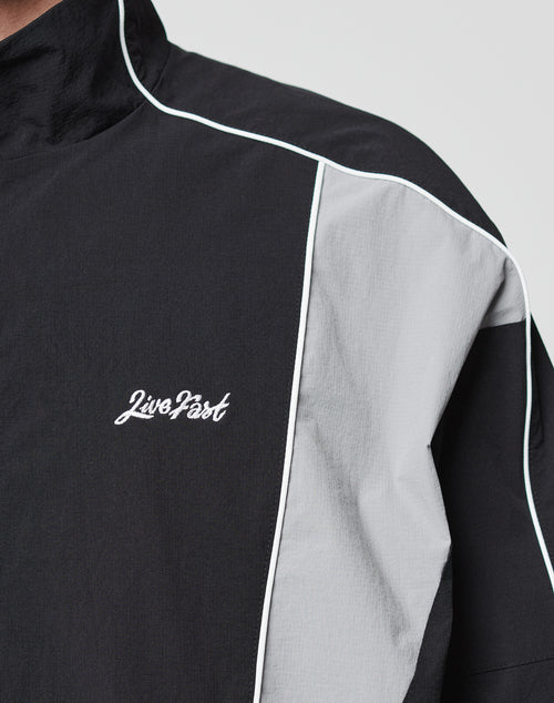 A close-up of the LIVE FAST DIE YOUNG Basic Nylon Trackjacket, a regular fit black jacket with white piping and a light gray panel, featuring “Live Fast” embroidered in white on the chest.