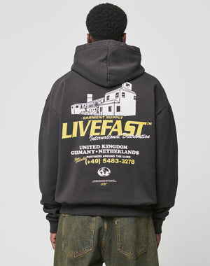 A person with short curly hair is shown from behind wearing the LIVE FAST DIE YOUNG LFDY Supply Hooded, an oversized black Baumwolle Polyester hoodie featuring bold white and yellow graphics, a building print, contact info, and country names.