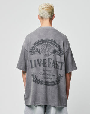 A person with short blond hair wears the LFDY Loyal Tee by LIVE FAST DIE YOUNG, a faded gray 100% Baumwolle oversize T-shirt with a large LIVE FAST graphic on the back, paired with light-wash denim shorts against a plain backdrop.