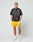 A man stands against a plain background wearing the oversized black LFDY Bark Tee by LIVE FAST DIE YOUNG, teamed with bright yellow shorts and white athletic shoes. His hands are in his pockets and he looks forward.