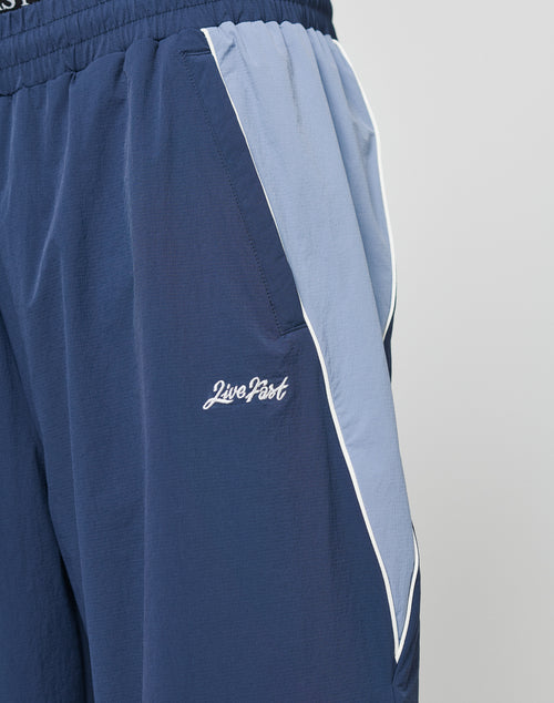 Close-up of a person wearing LIVE FAST DIE YOUNG Basic Nylon Trackpants, navy blue with elastic waistband, light blue and white pocket trim, zippered pockets, and the Live Fast logo embroidered near the pocket.