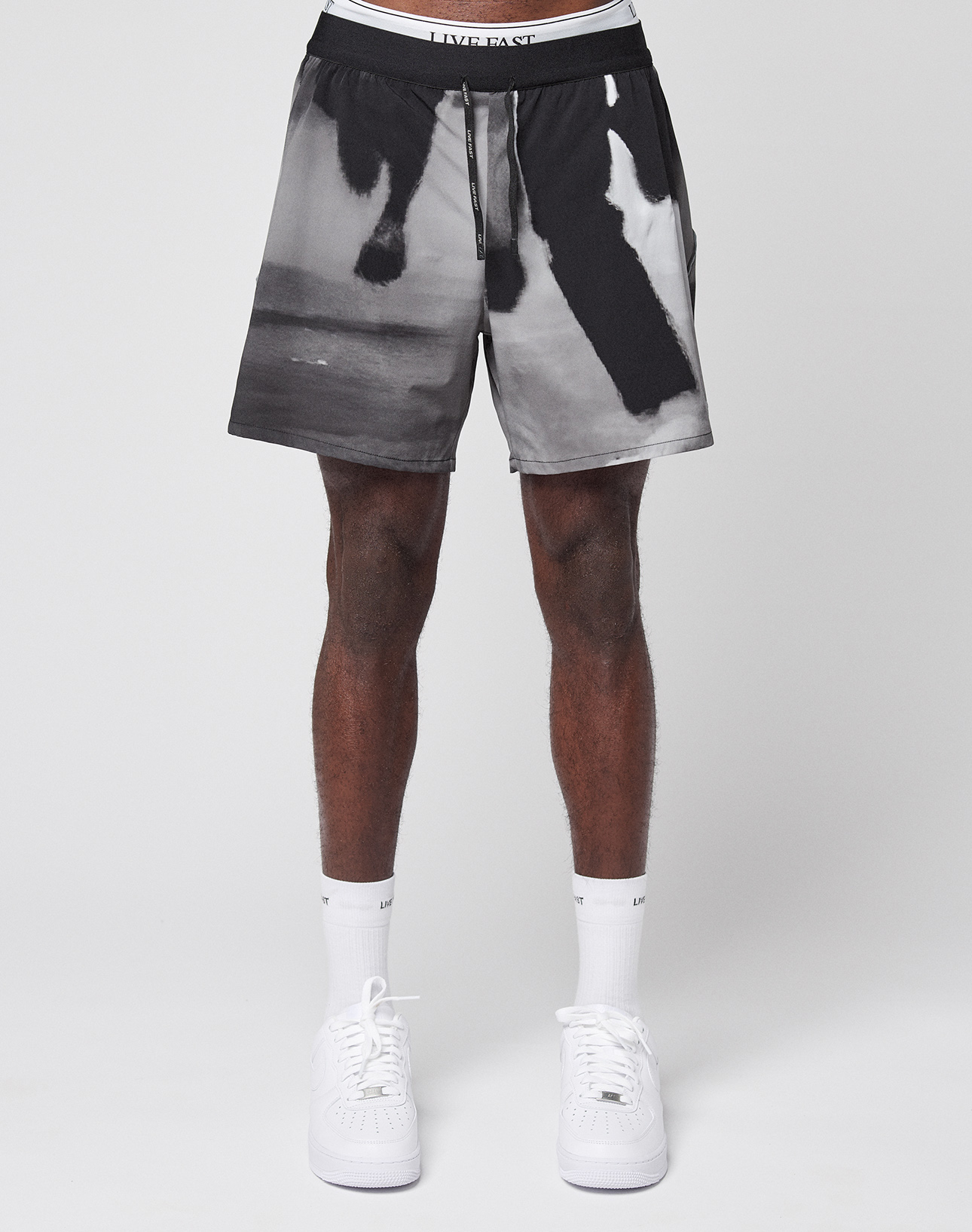 A person wears LFDY Performance Shorts by LIVE FAST DIE YOUNG, featuring a cat print and reflective logo, paired with white socks and sneakers. Only the lower half of their body is visible against a plain white background.