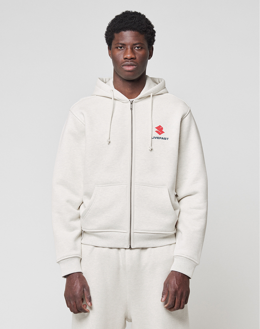 A person in a light beige LFDY Heavyweight Lounge Zip Hooded by LIVE FAST DIE YOUNG and matching sweatpants faces forward. The regular fit hoodie has a small red logo and “LIVEFAST” text on the left chest. Background is plain and light-colored.