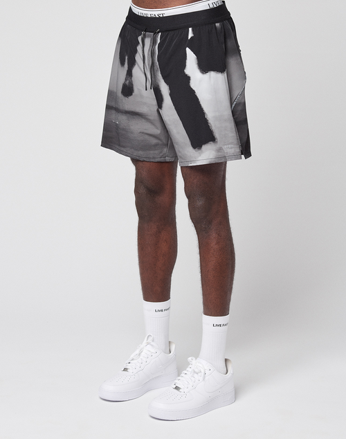 A person stands against a white background wearing LFDY Performance Shorts by LIVE FAST DIE YOUNG with a black-and-white abstract pattern, featuring a reflective logo print. White socks with LIVIN FAST text and white sneakers complete the cropped-below-waist image.