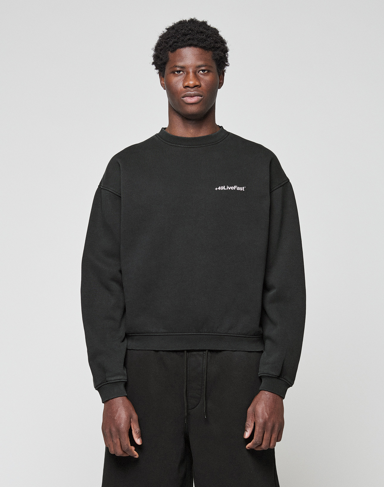 A young man with short curly hair wears the LIVE FAST DIE YOUNG Basic +49 Chest Sweater in black and black pants, standing against a plain light gray background. The sweater features small white #LiveFast text on the left chest.