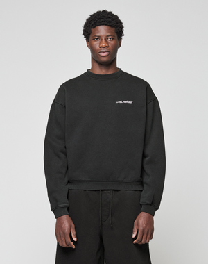 A young man with short curly hair wears the LIVE FAST DIE YOUNG Basic +49 Chest Sweater in black and black pants, standing against a plain light gray background. The sweater features small white #LiveFast text on the left chest.