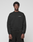 A young man with short curly hair wears the LIVE FAST DIE YOUNG Basic +49 Chest Sweater in black and black pants, standing against a plain light gray background. The sweater features small white #LiveFast text on the left chest.
