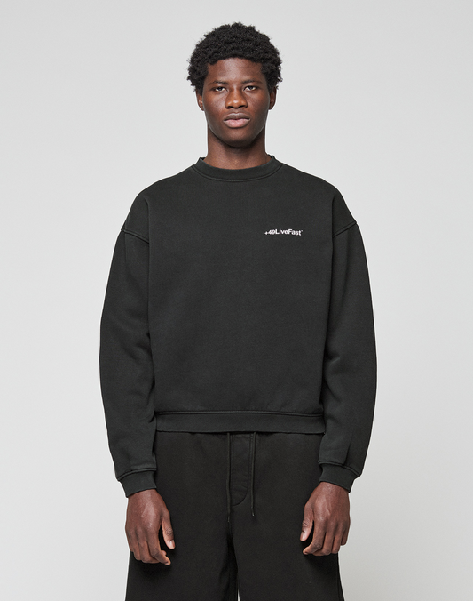 A young man with short curly hair wears the LIVE FAST DIE YOUNG Basic +49 Chest Sweater in black and black pants, standing against a plain light gray background. The sweater features small white #LiveFast text on the left chest.
