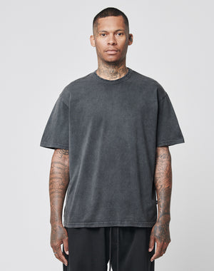 A tattooed person wears the LIVE FAST DIE YOUNG LFDY Blank Tee, a loose dark grey T-shirt aus 100% Baumwolle in Vintage-Waschung, and black regular fit pants, standing before a plain light grey background and looking straight ahead.