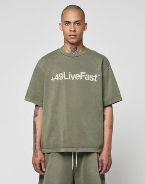 A person with a shaved head and tattoos wears a LIVE FAST DIE YOUNG Basic +49 Tee in olive green, made from 100% Baumwolle, featuring +49 LiveFast on the front, standing against a plain light background.