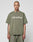 A person with a shaved head and tattoos wears a LIVE FAST DIE YOUNG Basic +49 Tee in olive green, made from 100% Baumwolle, featuring +49 LiveFast on the front, standing against a plain light background.