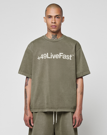 A person with a shaved head and visible tattoos wears the LIVE FAST DIE YOUNG Basic +49 Tee—an oversized olive-green shirt featuring a bold white "+49 Live Fast" front print—while standing against a plain light background.