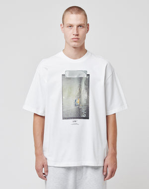 A person with short hair faces forward against a plain background, wearing the LIVE FAST DIE YOUNG Capri Tube Tee—an oversize white cotton t-shirt with a tape print graphic on the front—and light gray pants, with a neutral expression.