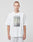 A person with short hair faces forward against a plain background, wearing the LIVE FAST DIE YOUNG Capri Tube Tee—an oversize white cotton t-shirt with a tape print graphic on the front—and light gray pants, with a neutral expression.