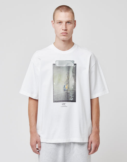 A person with short hair faces forward against a plain background, wearing the LIVE FAST DIE YOUNG Capri Tube Tee—an oversize white cotton t-shirt with a tape print graphic on the front—and light gray pants, with a neutral expression.