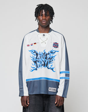 A man with short dreadlocks wears the LIVE FAST DIE YOUNG LFDY Flames Jersey—an oversized white and gray hockey top with blue flame graphics, front print details, and stripe patches—standing against a plain light background.