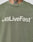 Someone wears an olive green Basic +49 Tee by LIVE FAST DIE YOUNG, featuring +49LiveFast™ in bold white on the chest and topstitching at the neckline. Part of a neck tattoo shows above the collar.