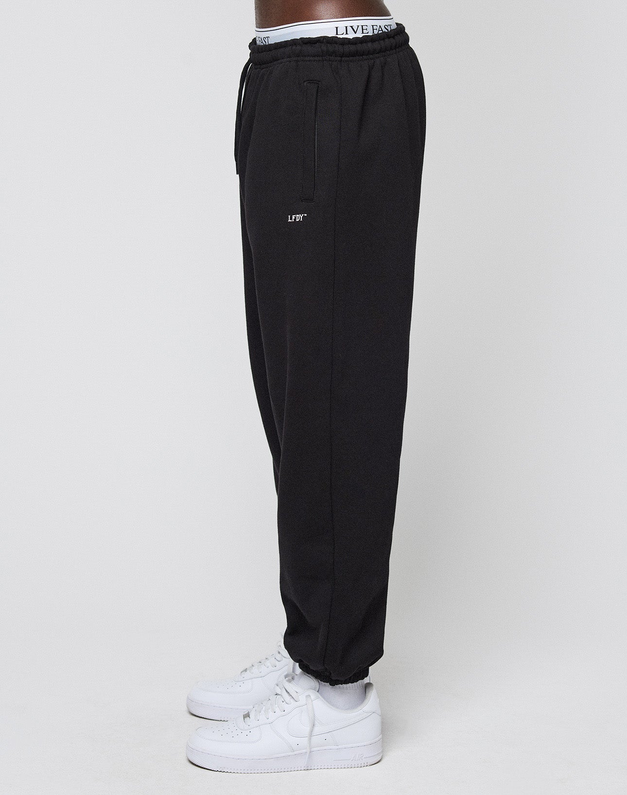 A person wearing LIVE FAST DIE YOUNG Basic Loose Jogger pants with elastic waistband and LFT logo on the thigh, paired with white sneakers, stands sideways against a plain white background.
