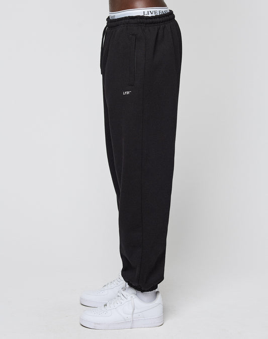 A person stands sideways in white sneakers against a plain background, wearing the LIVE FAST DIE YOUNG Basic Loose Jogger with logo print, an Elastik-Bund waistband reading LIVE FAST, and LIFT on the thigh.