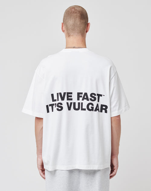 Someone faces away from the camera in a plain setting, wearing the LIVE FAST DIE YOUNG Capri Tube Tee—an oversized white cotton shirt with bold black LIVE FAST ITS VULGAR tape print on the back.