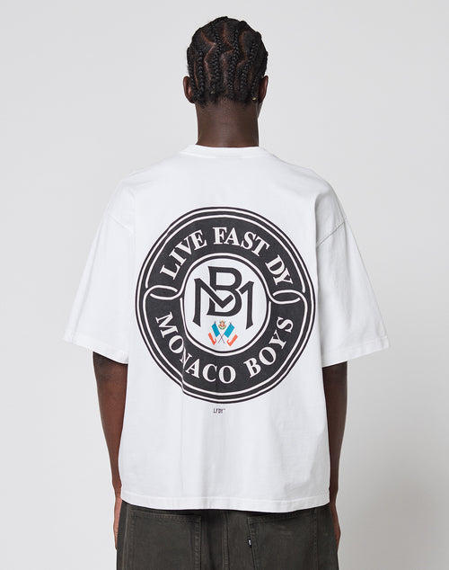 Seen from the back, a person with braided hair wears the LFDY Monaco Boys Tee by LIVE FAST DIE YOUNG—an oversized white 100% Baumwolle shirt with a large circular black graphic reading LIVE FAST BY MONACO BOYS and MB1 in the center.
