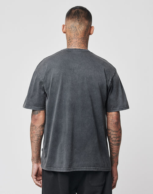 A tattooed man stands with his back to the camera, wearing a faded black LFDY Blank Tee by LIVE FAST DIE YOUNG in Vintage-Waschung and Regular Fit black pants, set against a plain light gray background.