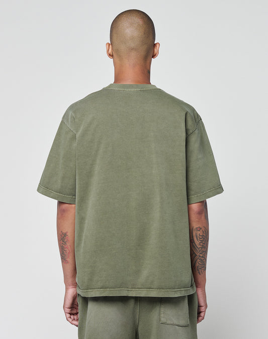A person with a shaved head stands with their back to the camera, wearing an olive green LIVE FAST DIE YOUNG Basic +49 Tee and matching 100% cotton shorts. Their tattooed arms hang loosely by their sides.