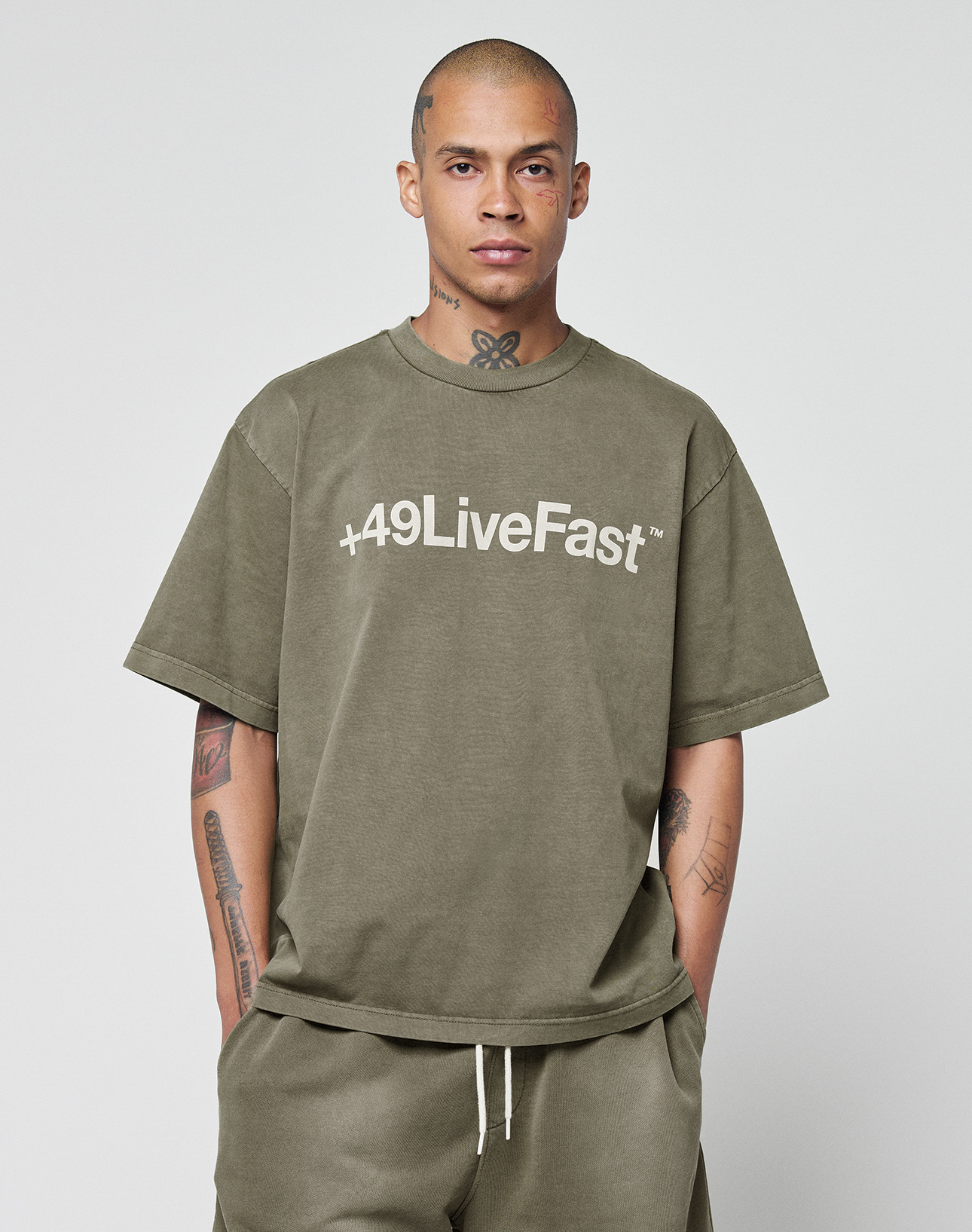 A person with a shaved head and tattoos wears the LIVE FAST DIE YOUNG Basic +49 Tee in olive green—an oversize fit t-shirt with bold frontprint—paired with matching shorts, both made from 100% Baumwolle, against a plain background.