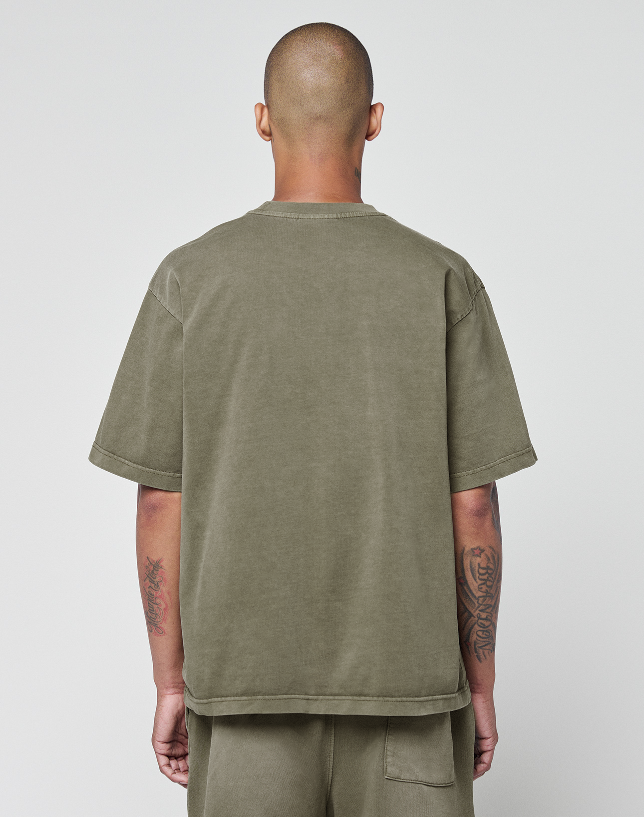 Seen from behind, a person with a shaved head wears the oversized Basic +49 Tee in olive green by LIVE FAST DIE YOUNG, paired with matching shorts. Tattooed arms add edge against a plain light backdrop.