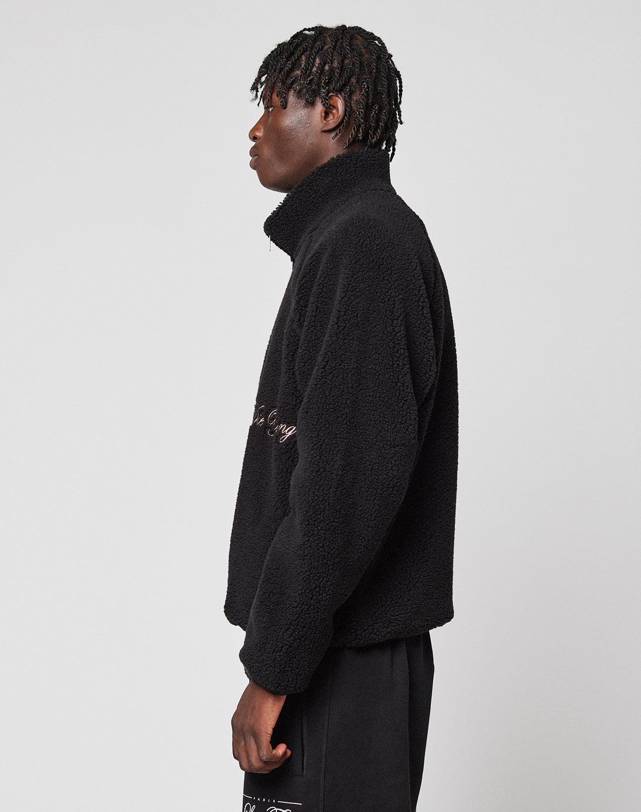 A person with short braided hair stands sideways in a schwarze LFDY Teddy Jacket von LIVE FAST DIE YOUNG und schwarzen Hosen vor einem schlichten, hellgrauen Hintergrund.