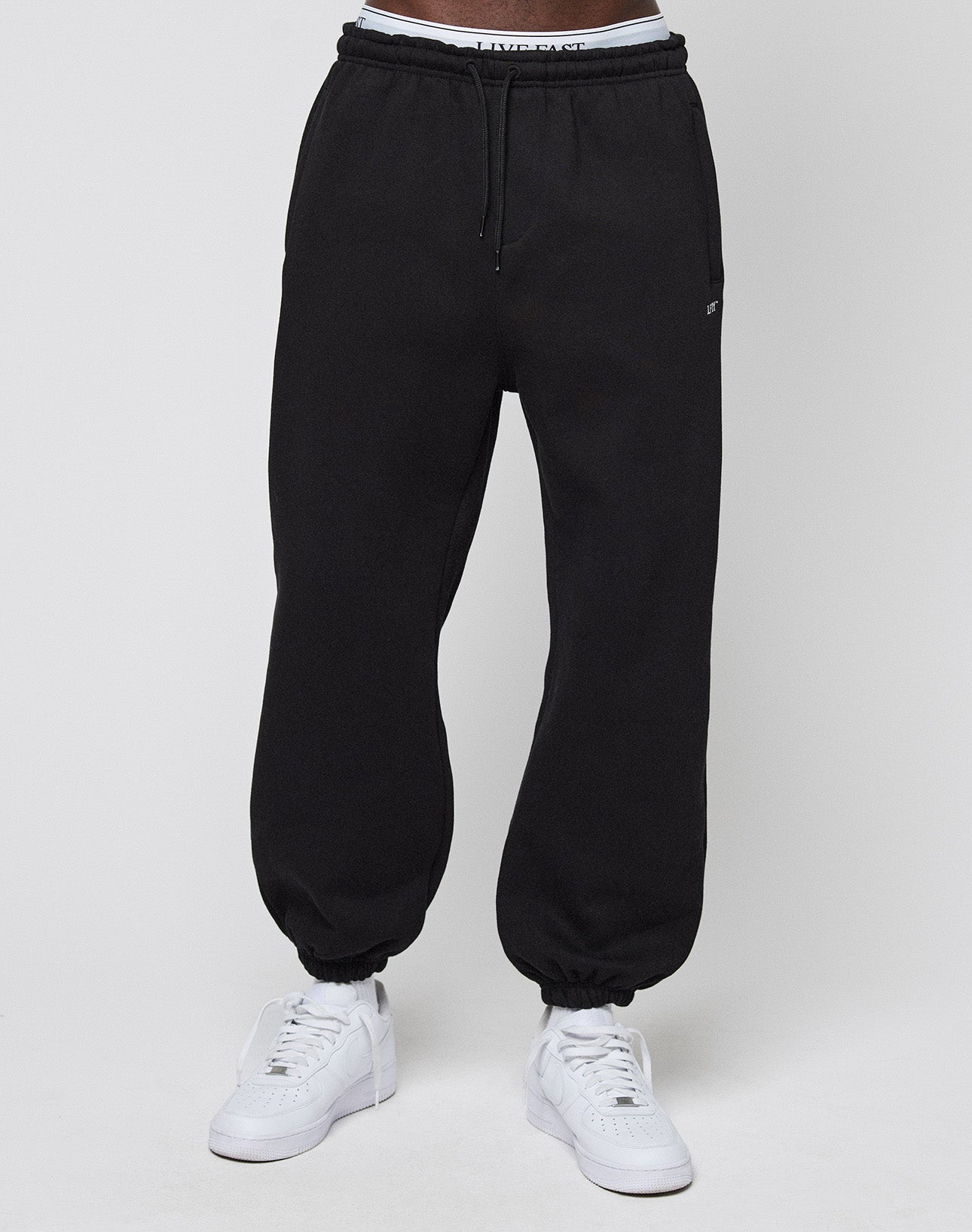 A person wears the LIVE FAST DIE YOUNG Basic Loose Jogger in black with an elastic waistband and cuffs, white sneakers, and slightly visible logo-print white underwear against a plain light gray background.
