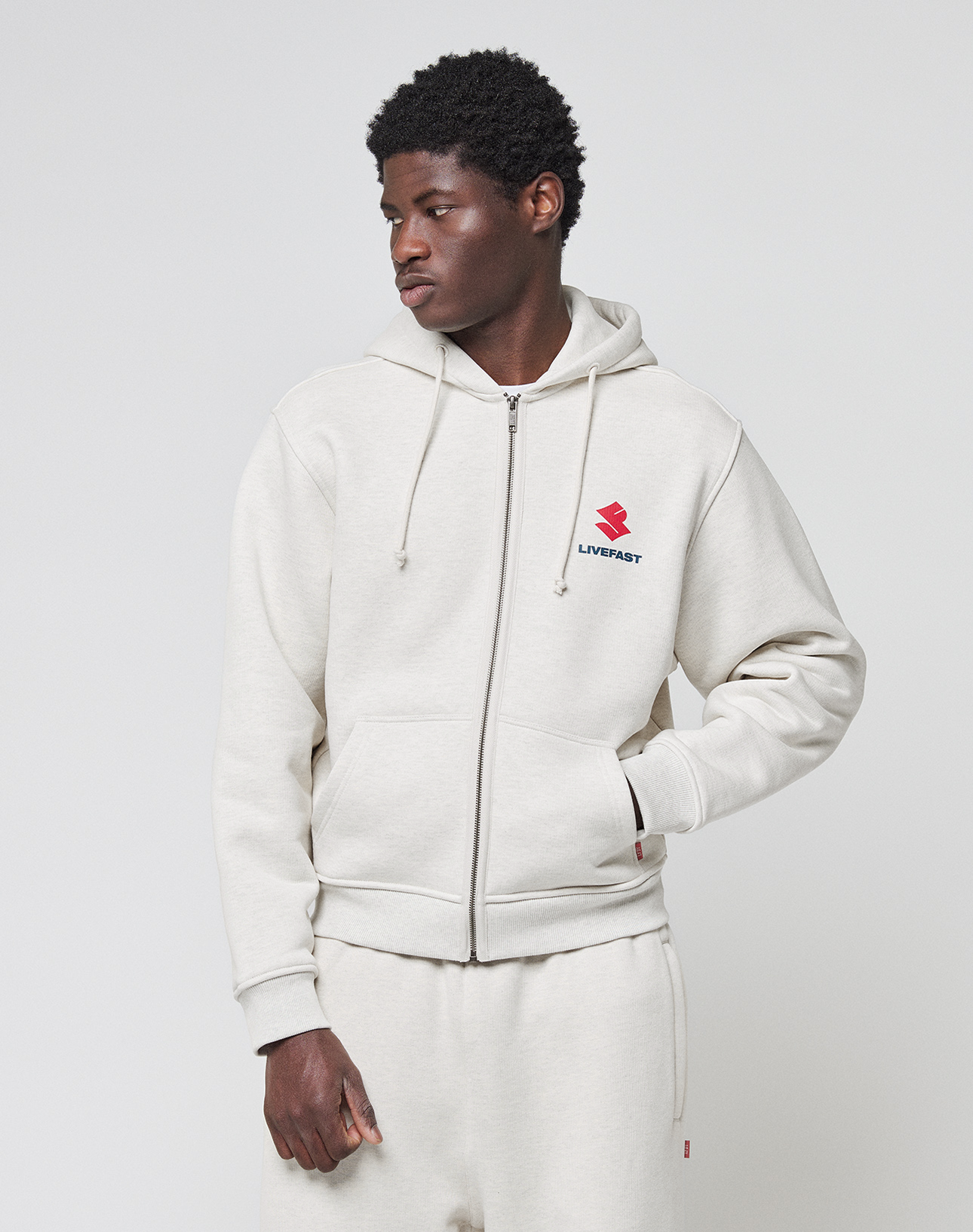 A young man wears a light beige LFDY Heavyweight Lounge Zip Hooded by LIVE FAST DIE YOUNG with matching sweatpants. The 100% cotton hoodie has a red and black LIVEFAST logo on the chest. He stands against a white background, looking to the side.
