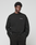 A man with short curly hair wears the LIVE FAST DIE YOUNG Basic +49 Chest Sweater in black, featuring a small white chest print, along with matching pants, and stands against a plain light gray background.