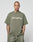A person with a shaved head and visible tattoos wears the Basic +49 Tee by LIVE FAST DIE YOUNG, an oversized olive-green 100% Baumwolle shirt with “+49LiveFast,” plus matching shorts, against a plain light background.