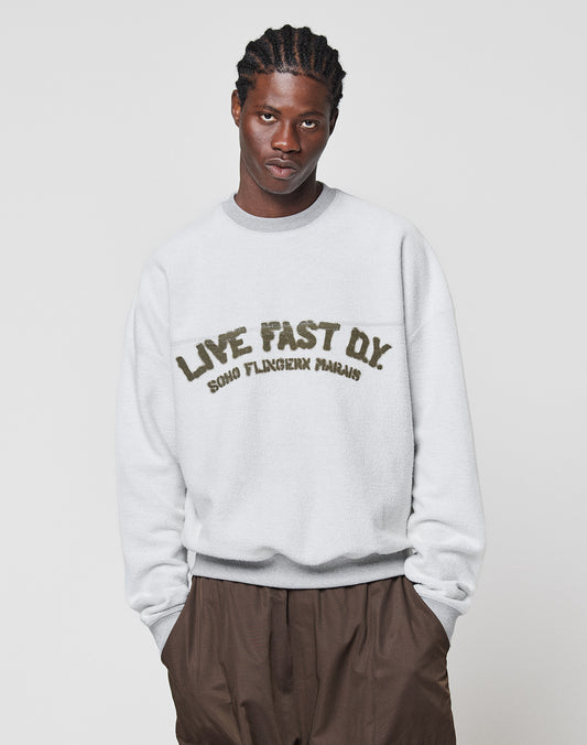 A man with braided hair wears the LFDY District Sweater by LIVE FAST DIE YOUNG, an oversized white Baumwolle-Polyester sweatshirt with bold LIVE FAST D.Y. SOHO FLAGSHIP PARADIS text, standing against a white background with hands in brown trousers pockets.