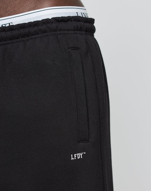 Close-up of a person wearing LIVE FAST DIE YOUNGs Basic Loose Jogger, featuring a black color, white LFDY™ logo by the pocket, elastic waistband, loose fit, and white underwear slightly visible above the pants.
