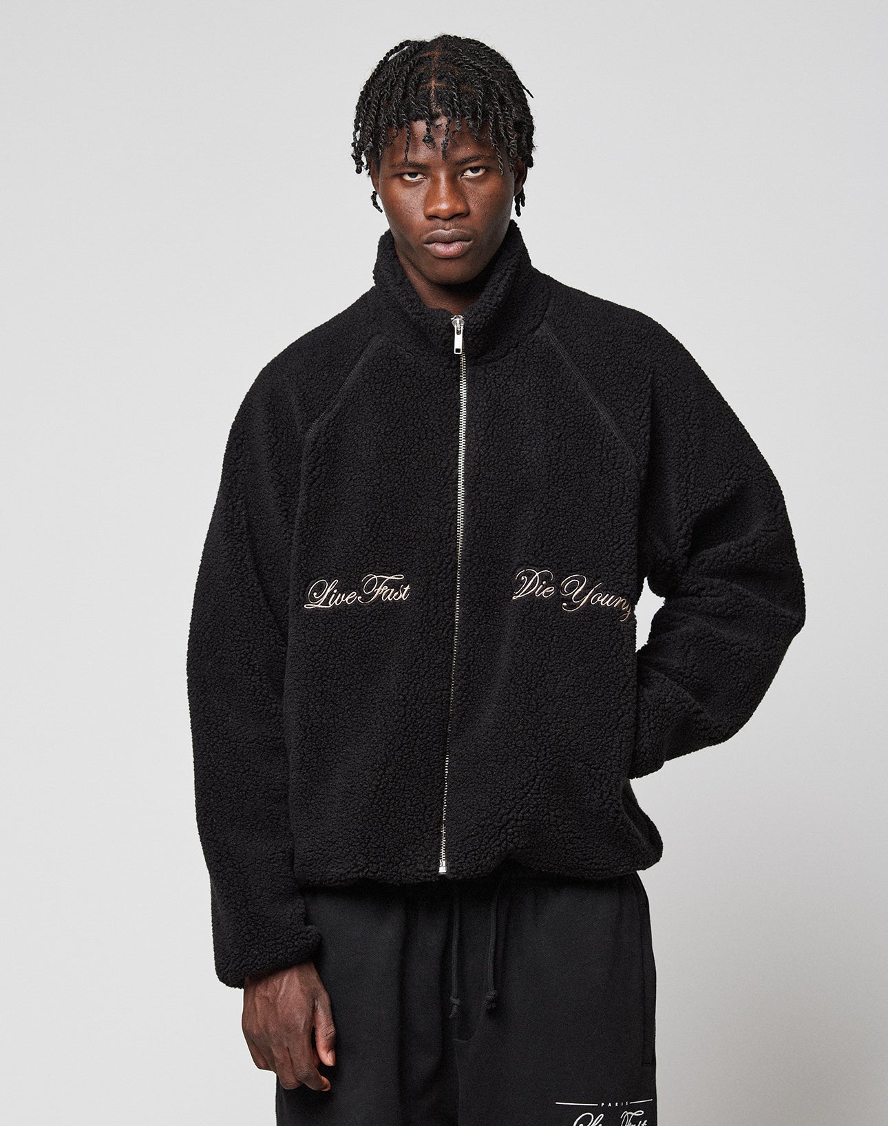 A person with short braided hair wears the LFDY Teddy Jacket by LIVE FAST DIE YOUNG, featuring raglan sleeves, a zipper, and white "Live Fast" and "Die Young" embroidery, paired with black pants against a plain light gray background.