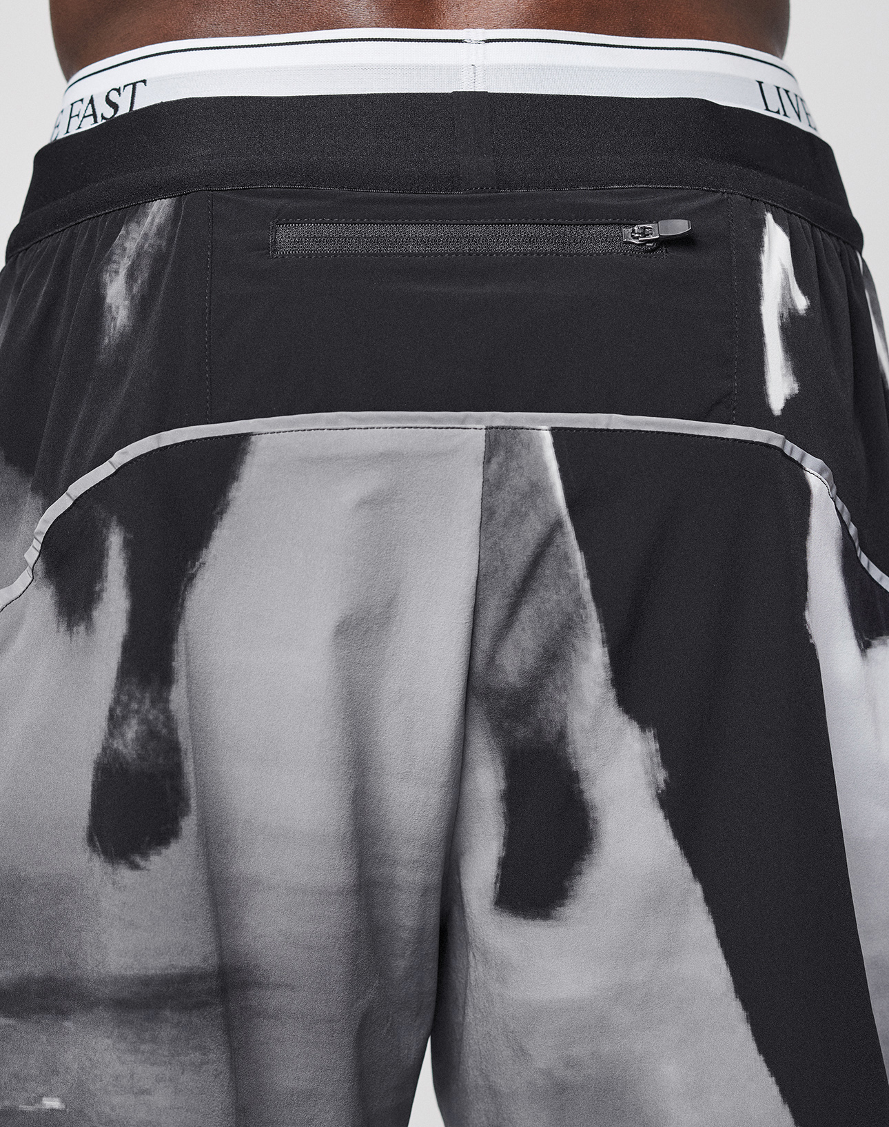 A close-up of the back of a person wearing LFDY Performance Shorts by LIVE FAST DIE YOUNG, featuring black and white abstract or animal print, a zipper pocket, "LIVE FAST" on the waistband, and a reflective logo print.