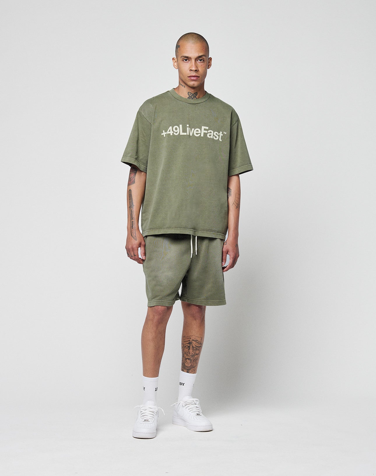 A person stands against a white background wearing the Basic +49 Tee in olive from LIVE FAST DIE YOUNG with topstitching at the neckline, matching shorts, white socks, sneakers, and several visible tattoos.