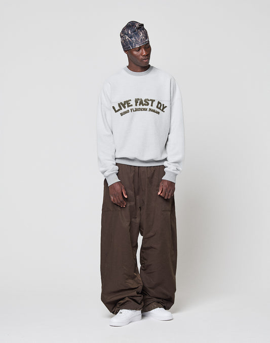 A man stands against a white background wearing a patterned headscarf, loose brown pants, white sneakers, and an oversized light gray LIVE FAST DIE YOUNG LFDY District Sweater.