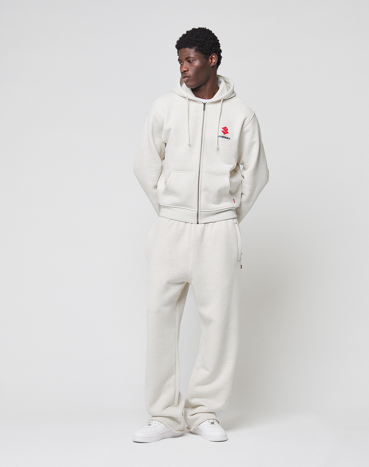 A man stands against a white background wearing LIVE FAST DIE YOUNG's off-white Heavyweight Loungepants and matching 650gsm hoodie with a red chest graphic, paired with white sneakers. His hands are in his pockets as he looks to the side.