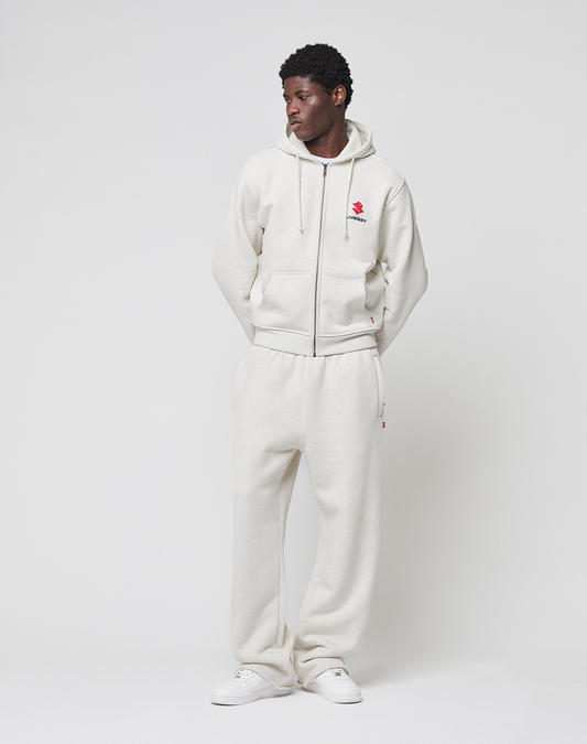 A man stands against a white background wearing the LIVE FAST DIE YOUNG LFDY Heavyweight Lounge Zip Hooded in light beige and matching sweatpants with white sneakers. His hands are in his pockets as he looks slightly to the side, highlighting the regular fit.
