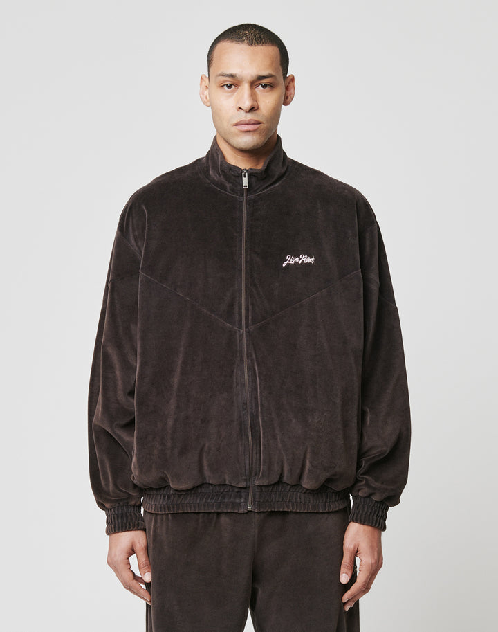 A person faces the camera in a LIVE FAST DIE YOUNG LFDY Velour Jacket, dark brown with an oversize fit, Stehkragen, zip-up front, and Lion Plan Logo-Stick embroidery on the left chest, paired with matching pants against a plain light background. | model