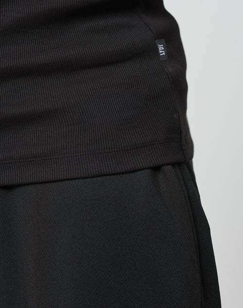 Close-up of a person wearing the LIVE FAST DIE YOUNG Blank Rib Tank, slim fit and ribbed in black, with a Flag Label on the side seam. The neutral background highlights the cotton texture and tank details.