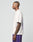 A man with short hair and arm tattoos stands in profile, wearing the oversize fit white LFDY Originals Tee from LIVE FAST DIE YOUNG and purple shorts against a plain light gray background.
