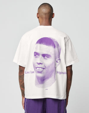 A person stands with their back to the camera wearing the LFDY Originals Tee by LIVE FAST DIE YOUNG—an oversized white cotton T-shirt featuring a large purple portrait backprint and Live Fast Originals text beside the image.