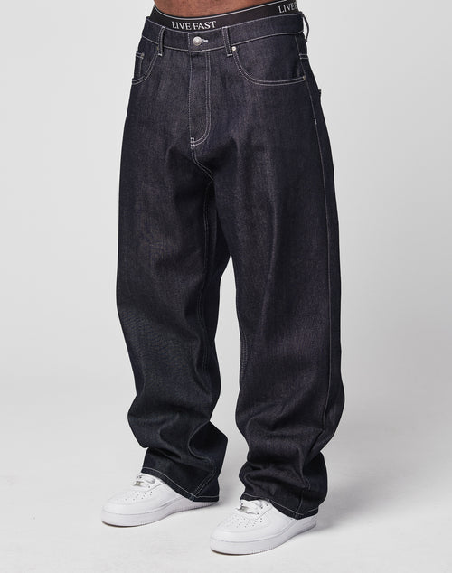 A person wears LFDY Baggy Denim jeans made from 100% cotton with a wide straight leg, white stitching, and a black waistband reading LIVE FAST. Theyre paired with white sneakers against a plain, light background.