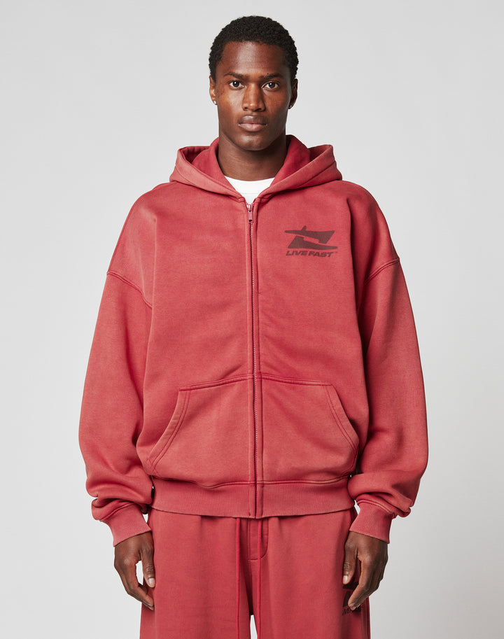 A man wears the LIVE FAST DIE YOUNG Live Fast Sweatsuit Jacket, a loose vintage-wash red zip-up with chest graphic and branded puller, matching red sweatpants, and a white shirt, standing against a plain gray background. | model